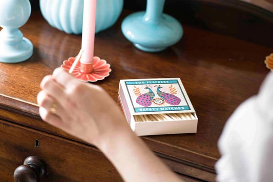 Caja de cerillas cuadrada con ilustración de dos pavos reales en azul y rosa sobre fondo crema, conteniendo cerillas largas de madera natural y punta blanca.