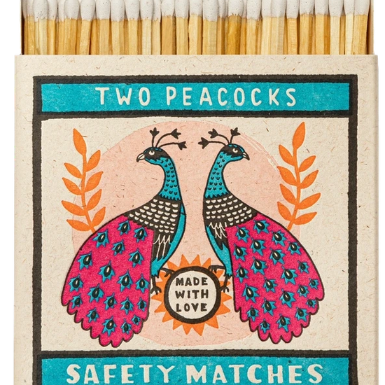 Caja de cerillas cuadrada con ilustración de dos pavos reales en azul y rosa sobre fondo crema, conteniendo cerillas largas de madera natural y punta blanca.