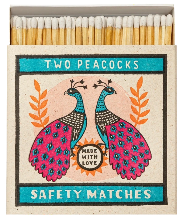 Caja de cerillas cuadrada con ilustración de dos pavos reales en azul y rosa sobre fondo crema, conteniendo cerillas largas de madera natural y punta blanca.