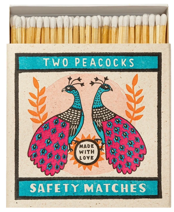 Caja de cerillas cuadrada con ilustración de dos pavos reales en azul y rosa sobre fondo crema, conteniendo cerillas largas de madera natural y punta blanca.