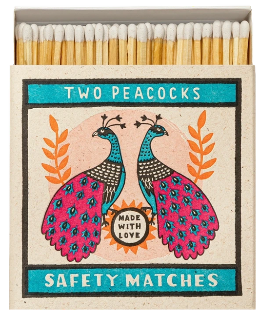 Caja de cerillas cuadrada con ilustración de dos pavos reales en azul y rosa sobre fondo crema, conteniendo cerillas largas de madera natural y punta blanca.