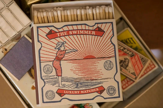 Caja de cerillas cuadrada con ilustración vintage de una mujer con bañador de rayas rojas y blancas saltando al mar, con cerillas largas de madera y punta blanca.