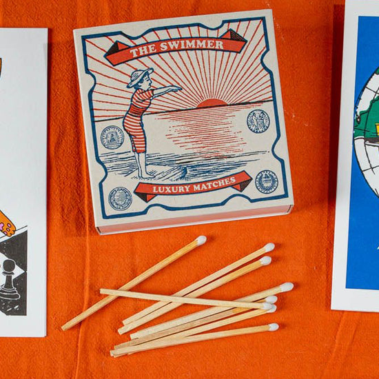 Caja de cerillas cuadrada con ilustración vintage de una mujer con bañador de rayas rojas y blancas saltando al mar, con cerillas largas de madera y punta blanca.