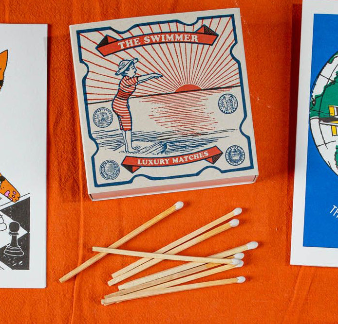 Caja de cerillas cuadrada con ilustración vintage de una mujer con bañador de rayas rojas y blancas saltando al mar, con cerillas largas de madera y punta blanca.