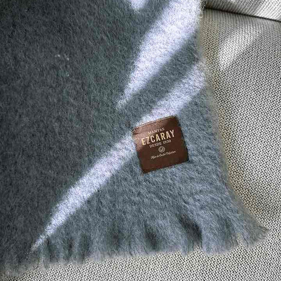 Manta de sofá de color azul grisáceo con textura de mohair esponjosa y brillante, terminada con flecos y fabricada de forma artesanal en España.