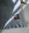 Manta de sofá de color azul grisáceo con textura de mohair esponjosa y brillante, terminada con flecos y fabricada de forma artesanal en España.