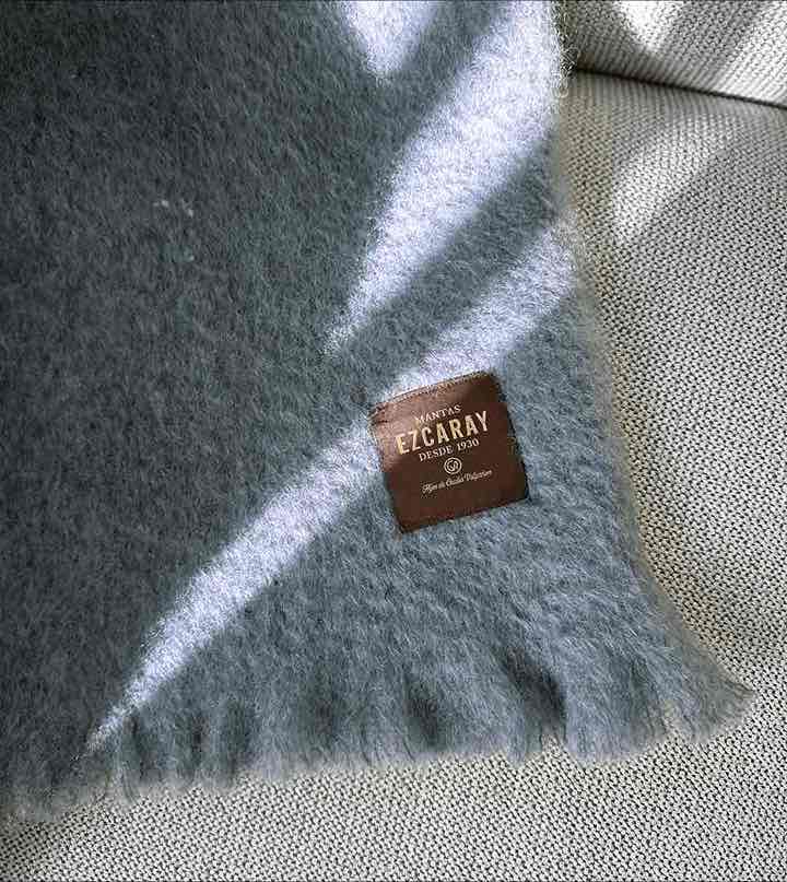 Manta de sofá de color azul grisáceo con textura de mohair esponjosa y brillante, terminada con flecos y fabricada de forma artesanal en España.