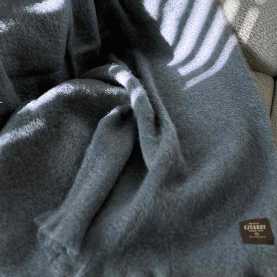 Manta de sofá de color azul grisáceo con textura de mohair esponjosa y brillante, terminada con flecos y fabricada de forma artesanal en España.