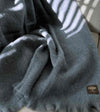 Manta de sofá de color azul grisáceo con textura de mohair esponjosa y brillante, terminada con flecos y fabricada de forma artesanal en España.