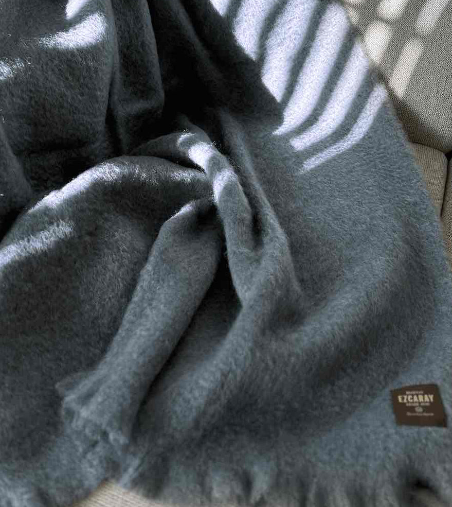 Manta de sofá de color azul grisáceo con textura de mohair esponjosa y brillante, terminada con flecos y fabricada de forma artesanal en España.