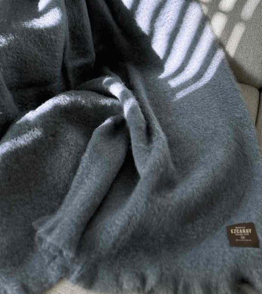Manta de sofá de color azul grisáceo con textura de mohair esponjosa y brillante, terminada con flecos y fabricada de forma artesanal en España.