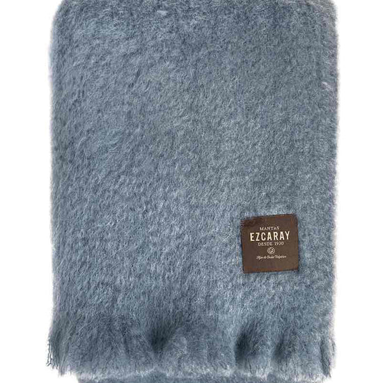 Manta de sofá de color azul grisáceo con textura de mohair esponjosa y brillante, terminada con flecos y fabricada de forma artesanal en España.