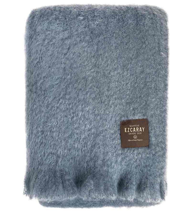 Manta de sofá de color azul grisáceo con textura de mohair esponjosa y brillante, terminada con flecos y fabricada de forma artesanal en España.