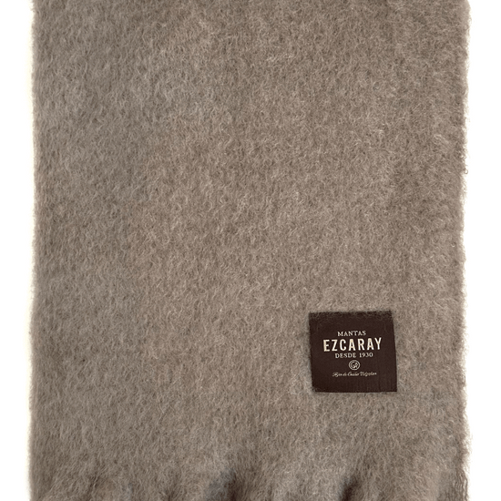 Manta de sofá de color topo con textura de mohair brillante y esponjosa, terminada en flecos manuales y fabricada mediante técnicas tradicionales en Ezcaray.