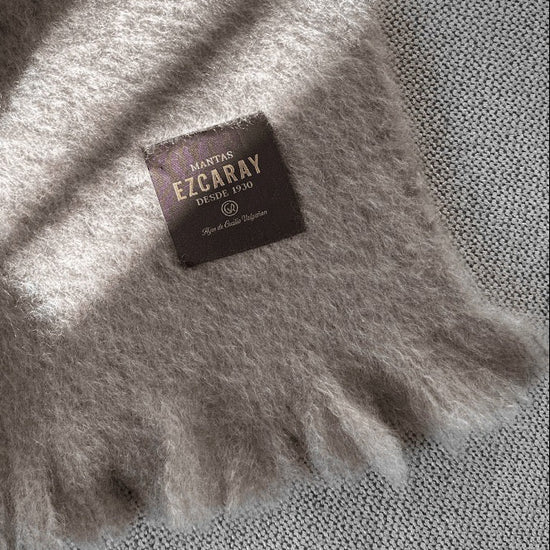 Manta de sofá de color topo con textura de mohair brillante y esponjosa, terminada en flecos manuales y fabricada mediante técnicas tradicionales en Ezcaray.