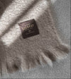 Manta de sofá de color topo con textura de mohair brillante y esponjosa, terminada en flecos manuales y fabricada mediante técnicas tradicionales en Ezcaray.