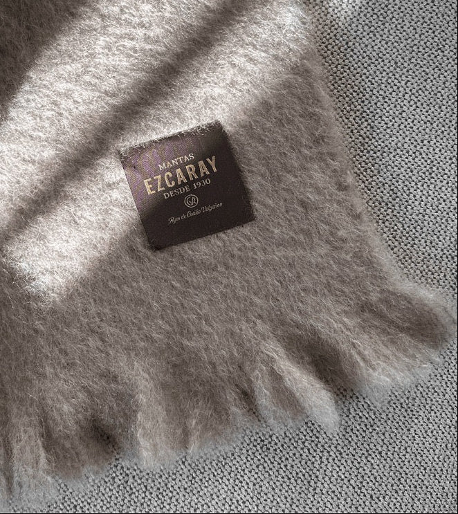 Manta de sofá de color topo con textura de mohair brillante y esponjosa, terminada en flecos manuales y fabricada mediante técnicas tradicionales en Ezcaray.