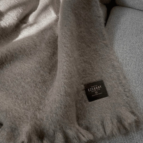 Manta de sofá de color topo con textura de mohair brillante y esponjosa, terminada en flecos manuales y fabricada mediante técnicas tradicionales en Ezcaray.