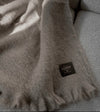 Manta de sofá de color topo con textura de mohair brillante y esponjosa, terminada en flecos manuales y fabricada mediante técnicas tradicionales en Ezcaray.