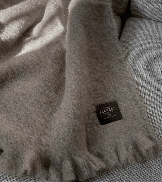 Manta de sofá de color topo con textura de mohair brillante y esponjosa, terminada en flecos manuales y fabricada mediante técnicas tradicionales en Ezcaray.