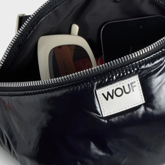 Riñonera negra con acabado brillante de la colección Nuit de Wouf, con correa ajustable, cremallera superior y etiqueta de cuero reciclado con logo frontal.