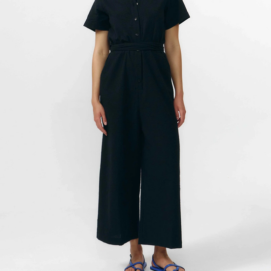 Mono largo de color negro con manga corta, cuello de camisa y pernera ancha, que incluye un cinturón a juego y botones naturales en la parte frontal.