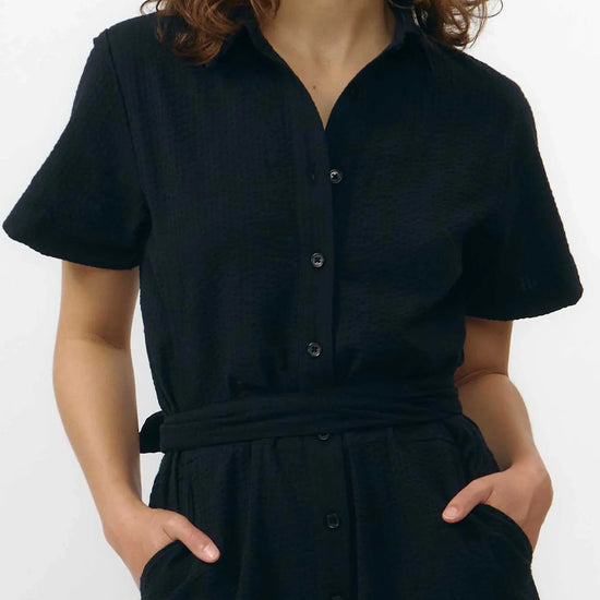 Mono largo de color negro con manga corta, cuello de camisa y pernera ancha, que incluye un cinturón a juego y botones naturales en la parte frontal.
