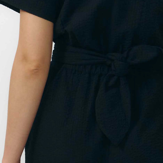 Mono largo de color negro con manga corta, cuello de camisa y pernera ancha, que incluye un cinturón a juego y botones naturales en la parte frontal.