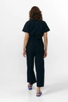Mono largo de color negro con manga corta, cuello de camisa y pernera ancha, que incluye un cinturón a juego y botones naturales en la parte frontal.