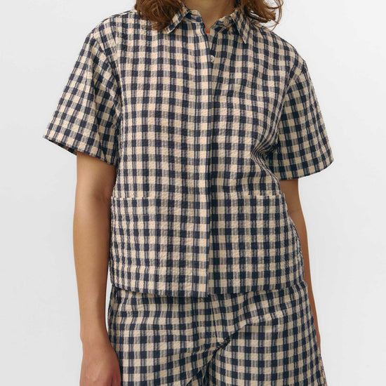 Camisa de manga corta con estampado de cuadros en tonos grises, cuello de solapa, dos bolsillos frontales cuadrados y cierre de botones naturales de corozo.