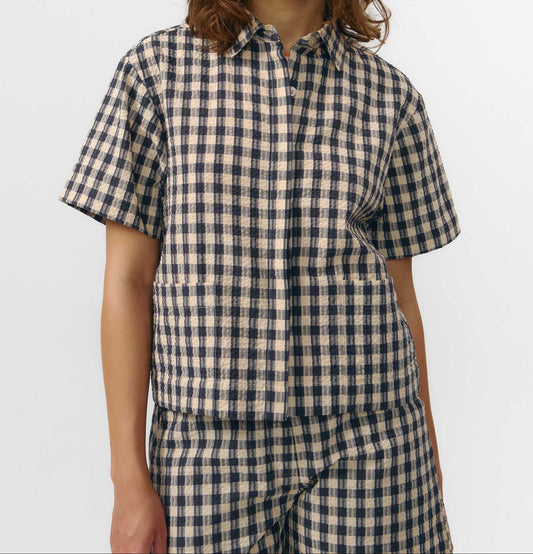 Camisa de manga corta con estampado de cuadros en tonos grises, cuello de solapa, dos bolsillos frontales cuadrados y cierre de botones naturales de corozo.