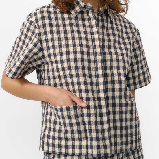 Camisa de manga corta con estampado de cuadros en tonos grises, cuello de solapa, dos bolsillos frontales cuadrados y cierre de botones naturales de corozo.