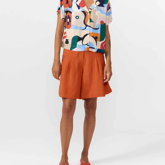 Camisa de manga corta con cuello de solapa y estampado artístico de barcos y motivos marineros en color azul sobre fondo crema, con botones de material natural.