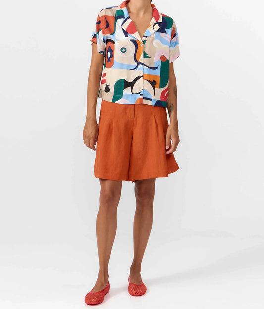 Camisa de manga corta con cuello de solapa y estampado artístico de barcos y motivos marineros en color azul sobre fondo crema, con botones de material natural.