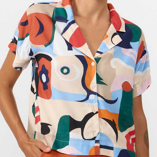 Camisa de manga corta con cuello de solapa y estampado artístico de barcos y motivos marineros en color azul sobre fondo crema, con botones de material natural.