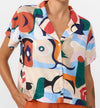 Camisa de manga corta con cuello de solapa y estampado artístico de barcos y motivos marineros en color azul sobre fondo crema, con botones de material natural.