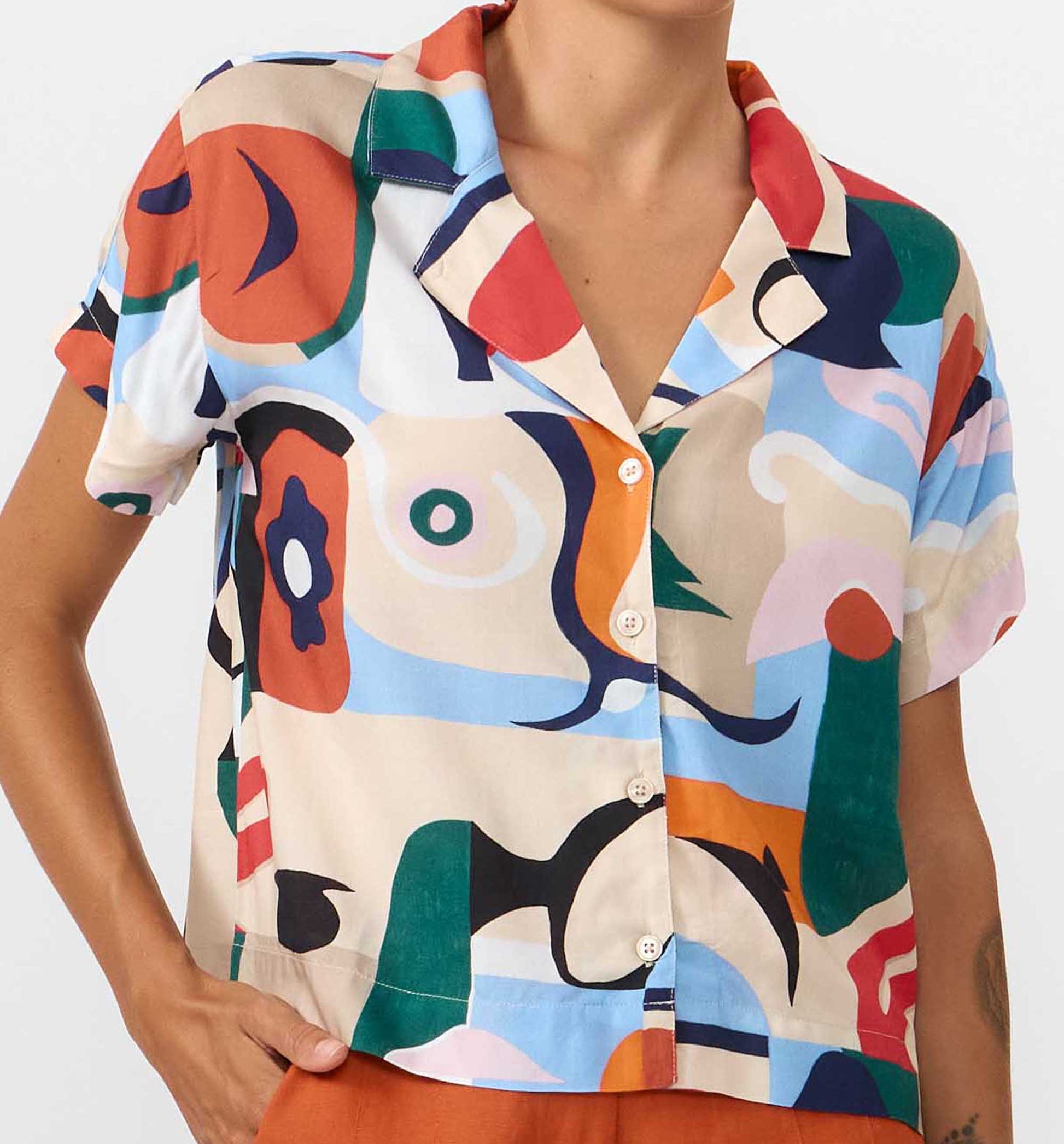 Camisa de manga corta con cuello de solapa y estampado artístico de barcos y motivos marineros en color azul sobre fondo crema, con botones de material natural.