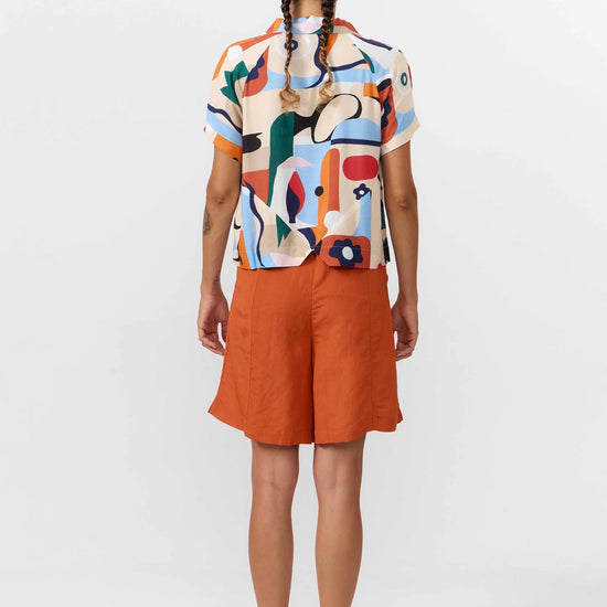 Camisa de manga corta con cuello de solapa y estampado artístico de barcos y motivos marineros en color azul sobre fondo crema, con botones de material natural.